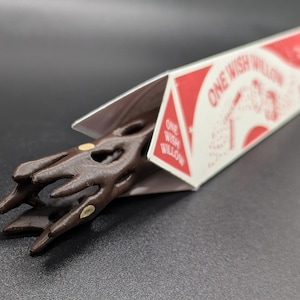 May include: A dark brown, carved wooden wand with a textured, branch-like handle featuring small, light-colored circular inlays. The wand is partially encased in a white triangular cardboard box labeled with the red text ONE WISH WILLOW.