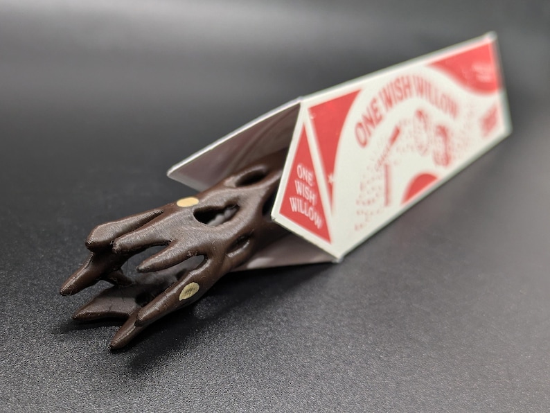 May include: A dark brown, carved wooden wand with a textured, branch-like handle featuring small, light-colored circular inlays. The wand is partially encased in a white triangular cardboard box labeled with the red text ONE WISH WILLOW.