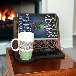May include: A wooden book stand with a decorative cutout design holds a copy of "The Ultimate Hitchhiker's Guide to the Galaxy" by Douglas Adams. A mug with a sheep design and a pair of black glasses are on the stand. The stand is on a wooden table.