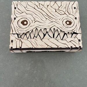 May include: A wooden box with a carved monster face. The box is made of light wood and has a dark brown stain. The monster has two large eyes and a wide, toothy grin.