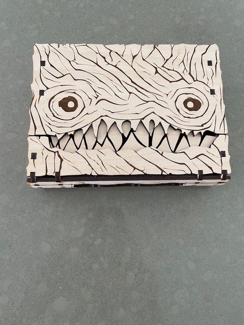 May include: A wooden box with a carved monster face. The box is made of light wood and has a dark brown stain. The monster has two large eyes and a wide, toothy grin.