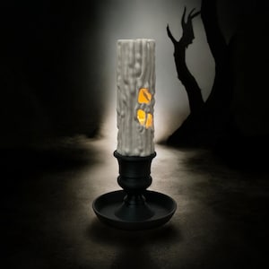 May include: A white candle with a carved design resembling a tree trunk, with a glowing orange light inside. The candle is on a black candlestick with a round base.