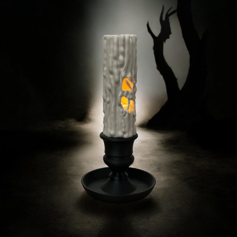 May include: A white candle with a carved design resembling a tree trunk, with a glowing orange light inside. The candle is on a black candlestick with a round base.