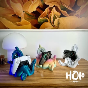 May include: Three 3D printed octopus-shaped game controller holders. Each holder is a different color: teal, white, and a gradient of pink and blue. The teal holder has a black and white PlayStation 5 controller, the white holder has a black Xbox controller, and the pink and blue holder has a black and green Nintendo Switch controller. The holders are on a wooden surface with the text "HOLO - PROPS" printed on the surface.