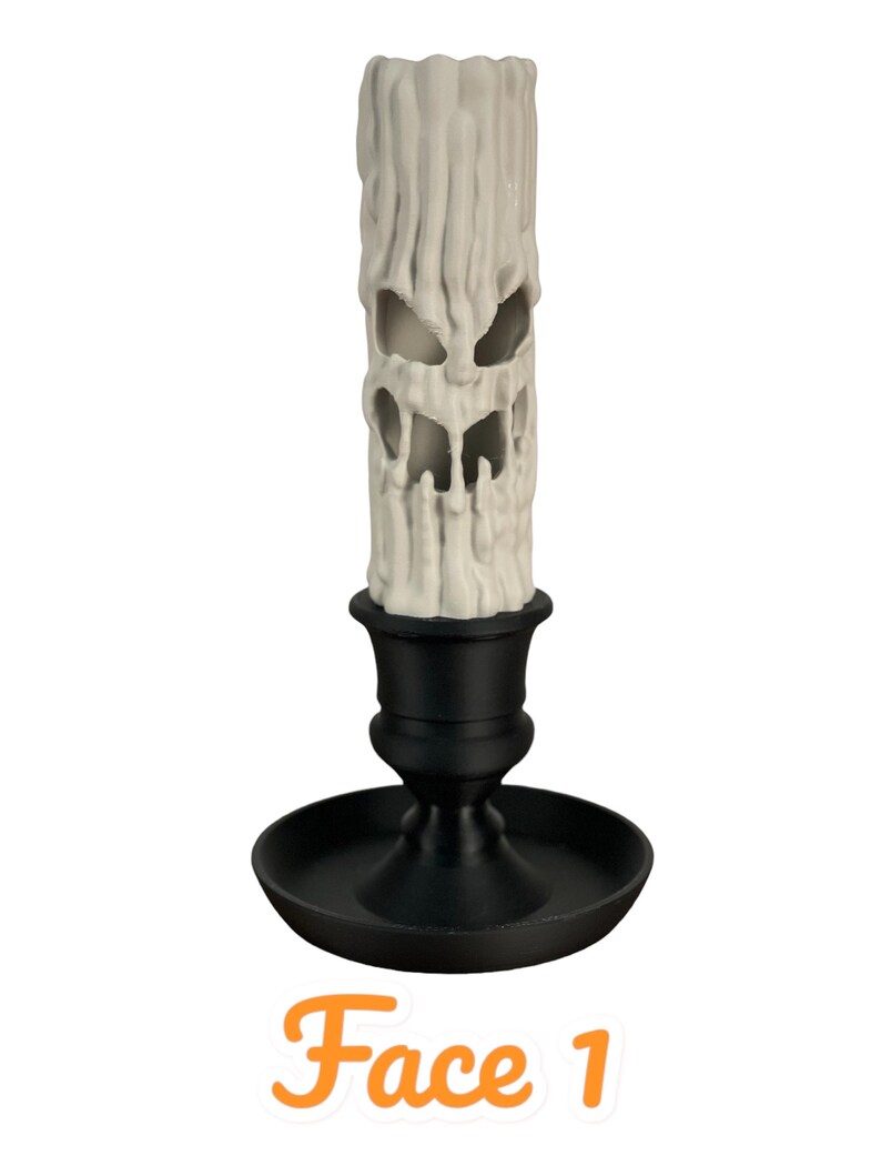 May include: A white candle with a carved face, sitting in a black candlestick. The candle is shaped like a melting face with a wide mouth and two eyes. The candlestick has a round base and a tall, slender stem. "Face 1" is written below the candle.