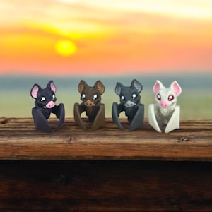 May include: Four small bat figurines with wings outstretched, sitting on a wooden surface. The bats are black, brown, gray, and white.