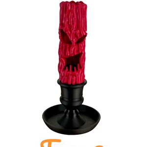 May include: A red candle with a carved face, resembling a jack-o'-lantern, is placed in a black candlestick. The candle is textured and has a menacing expression. The text "Face 2" is written below the candle.