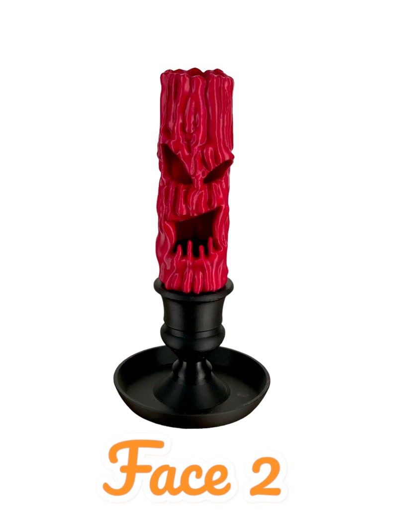 May include: A red candle with a carved face, resembling a jack-o'-lantern, is placed in a black candlestick. The candle is textured and has a menacing expression. The text "Face 2" is written below the candle.