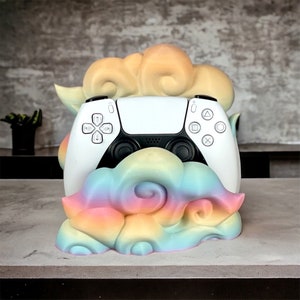May include: A white PlayStation 5 controller sits in a rainbow-colored cloud-shaped stand. The stand is made of a soft, textured material and has a whimsical design.