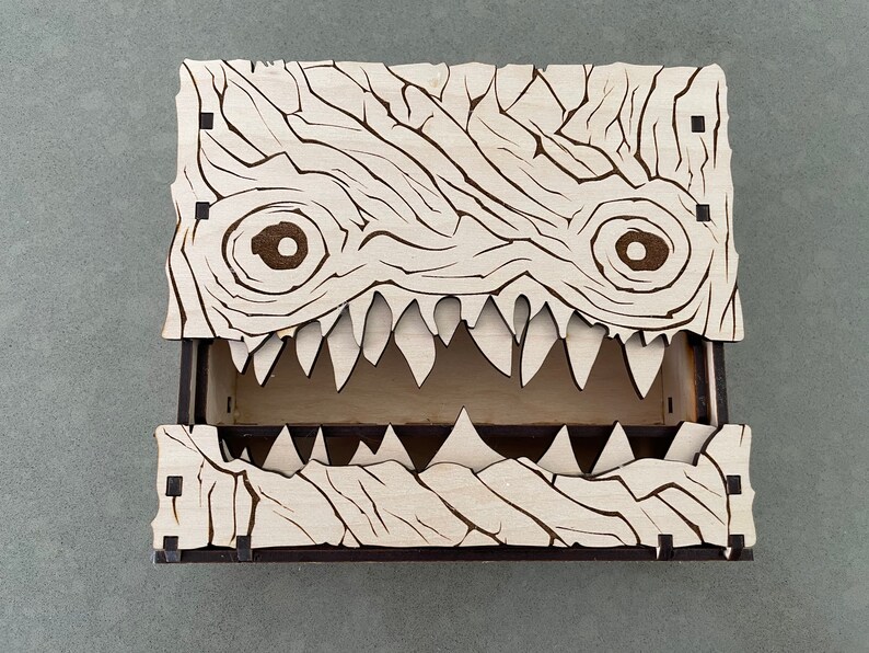 May include: A wooden box with a monster face design. The box has a hinged lid and a compartment inside. The monster face has two eyes, a mouth with sharp teeth, and a textured surface.