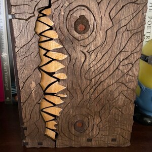 May include: A wooden book box with a carved design of a monster's face with two eyes and a mouth full of teeth. The box is made of dark brown wood with a light brown wood inlay for the teeth.