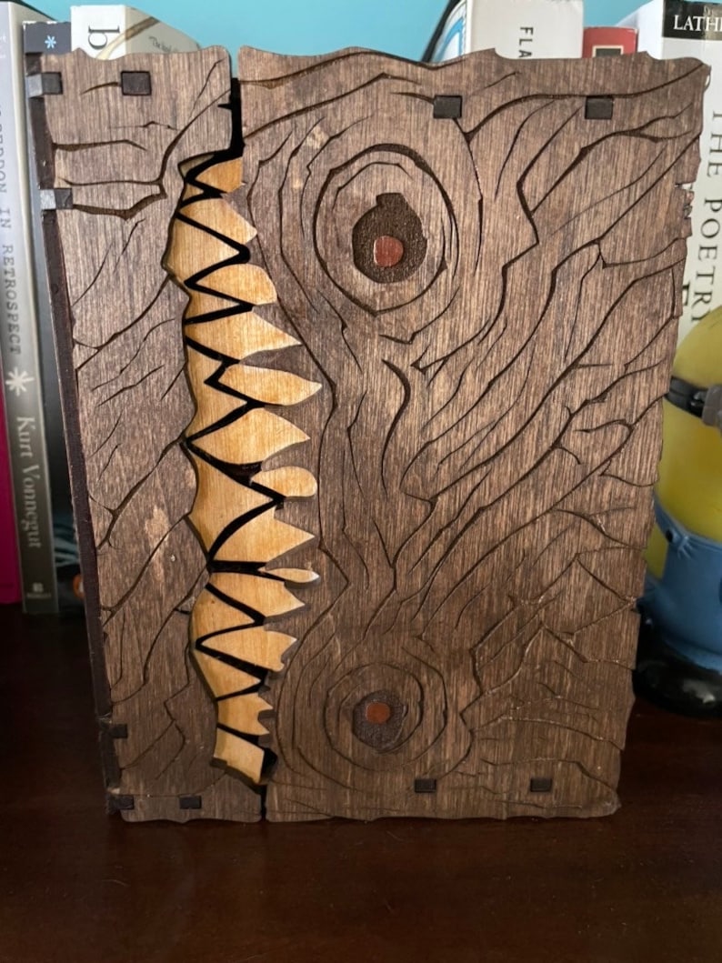 May include: A wooden book box with a carved design of a monster's face with two eyes and a mouth full of teeth. The box is made of dark brown wood with a light brown wood inlay for the teeth.