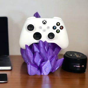 Op de afbeelding: Een witte Xbox-controller staat op een paarse, kristalvormige standaard. De controller heeft de standaard Xbox-knoppen, waaronder A, B, X, Y en een directionele pad. De standaard is gemaakt van 3D-geprint materiaal en heeft een getextureerd oppervlak.