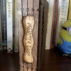 May include: A wooden box with a hinged lid, the front of which is engraved with the words "Monster Book". The box is brown with a light wood interior.