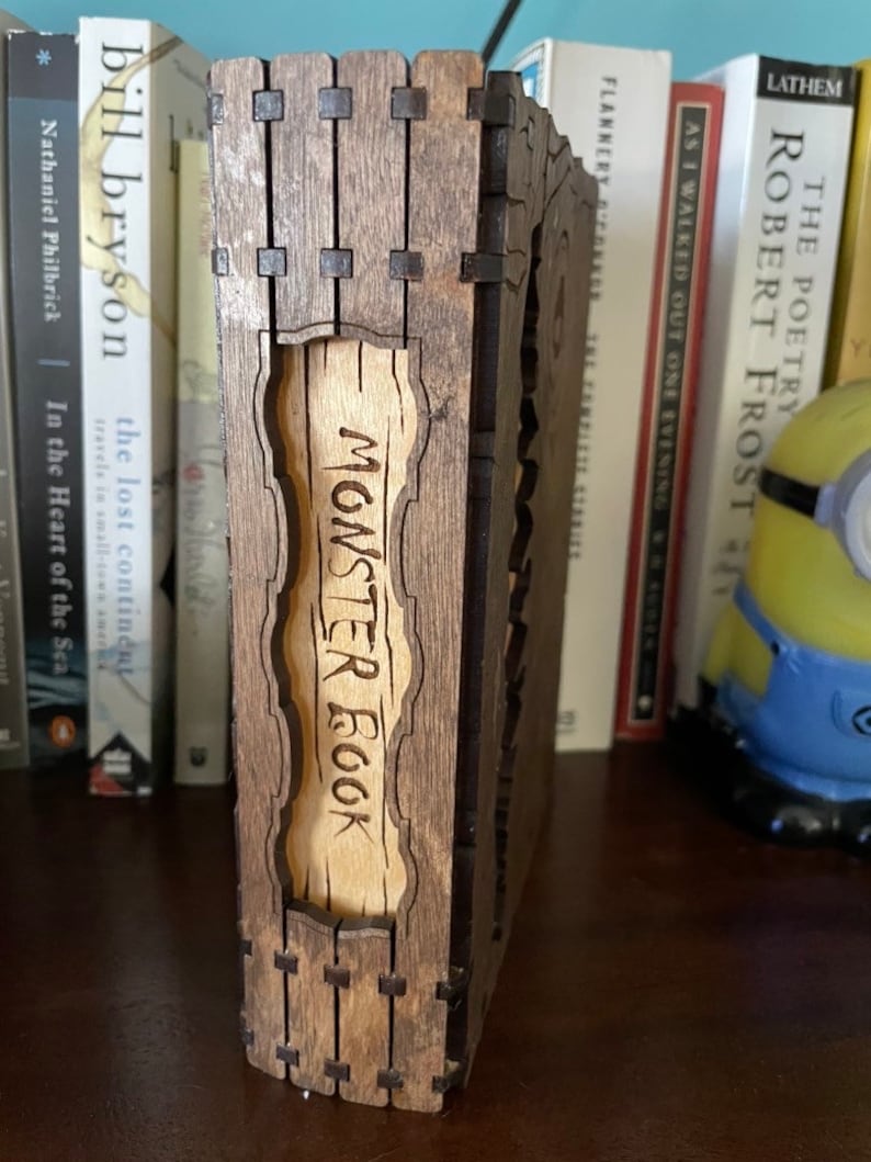 May include: A wooden box with a hinged lid, the front of which is engraved with the words "Monster Book". The box is brown with a light wood interior.