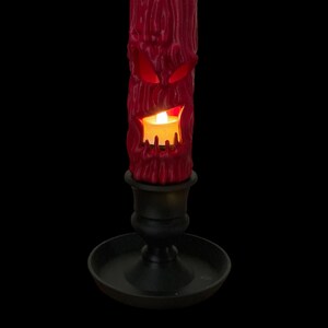 May include: A red candle with a carved face and a lit flame sits in a black candle holder.