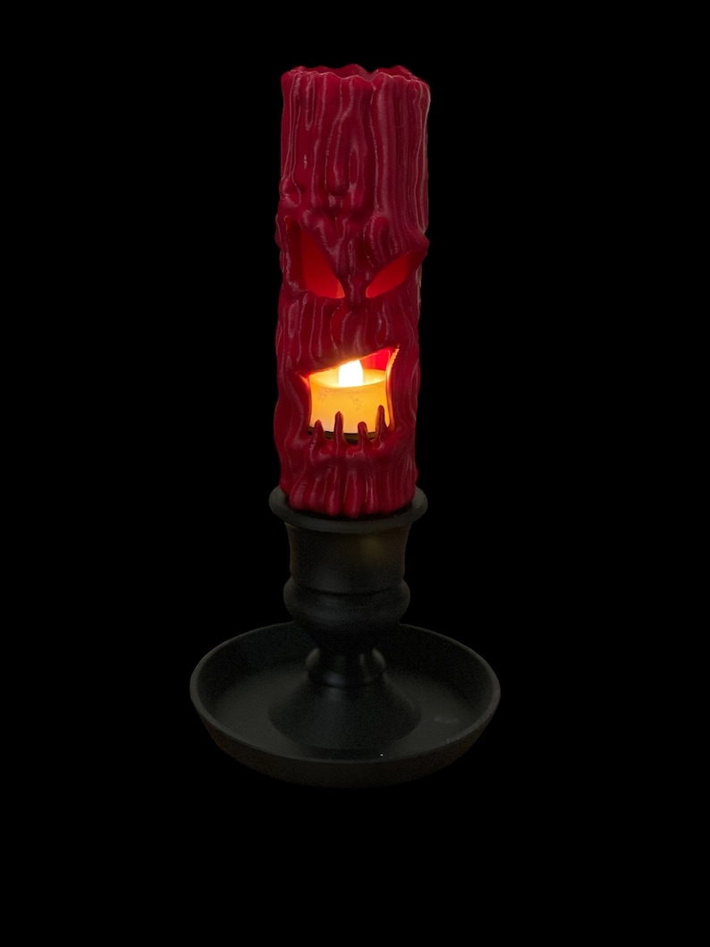May include: A red candle with a carved face and a lit flame sits in a black candle holder.