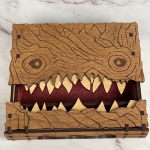 May include: Wooden dice box with a carved monster face design. The box has a hinged lid and a red felt interior. The monster's mouth is open, revealing rows of sharp teeth.