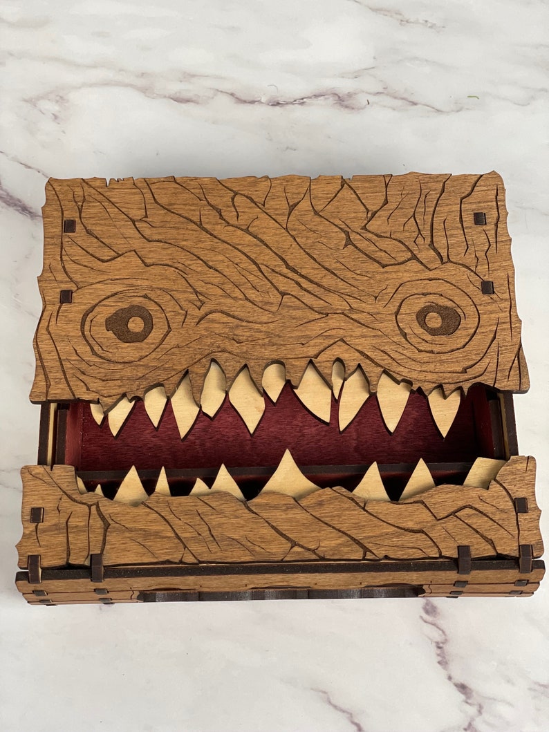 May include: Wooden dice box with a carved monster face design. The box has a hinged lid and a red felt interior. The monster's mouth is open, revealing rows of sharp teeth.