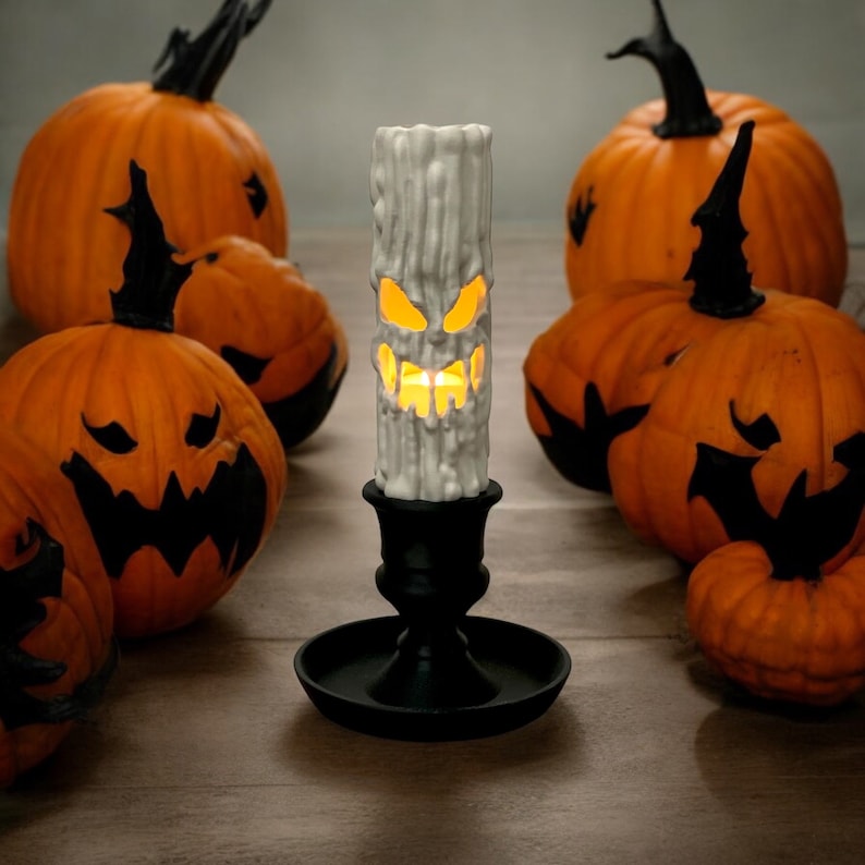 May include: A white candle with a carved jack-o'-lantern face sits on a black candlestick. The candle is surrounded by several orange pumpkins with black jack-o'-lantern faces.