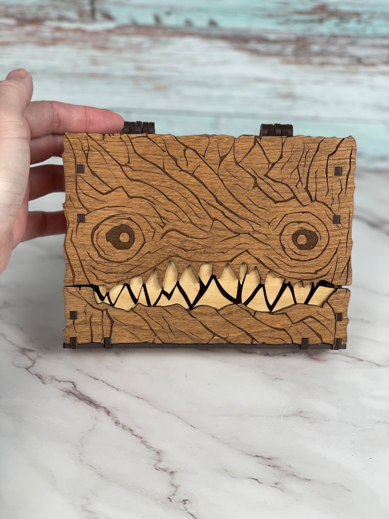 May include: A wooden box with a carved monster face. The box is made of light brown wood with dark brown accents. The monster has a wide, toothy grin and large, round eyes.