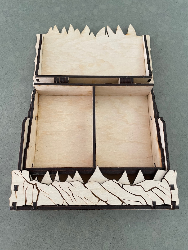 May include: A wooden box with a hinged lid and two compartments. The box is decorated with a design of jagged peaks, resembling a mountain range.