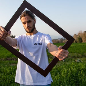 May include: A person wearing a white t-shirt with the text "INVOKA" in black, holding a dark brown square frame. The person is standing in a field of green grass under a blue sky.