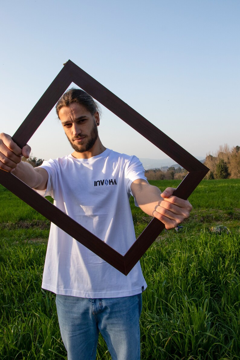 May include: A person wearing a white t-shirt with the text "INVOKA" in black, holding a dark brown square frame. The person is standing in a field of green grass under a blue sky.
