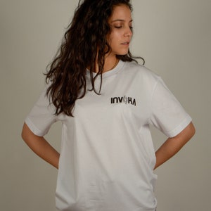 May include: A white t-shirt with a black graphic that says "INVIKA" on the front. The person wearing the shirt has long, curly brown hair.
