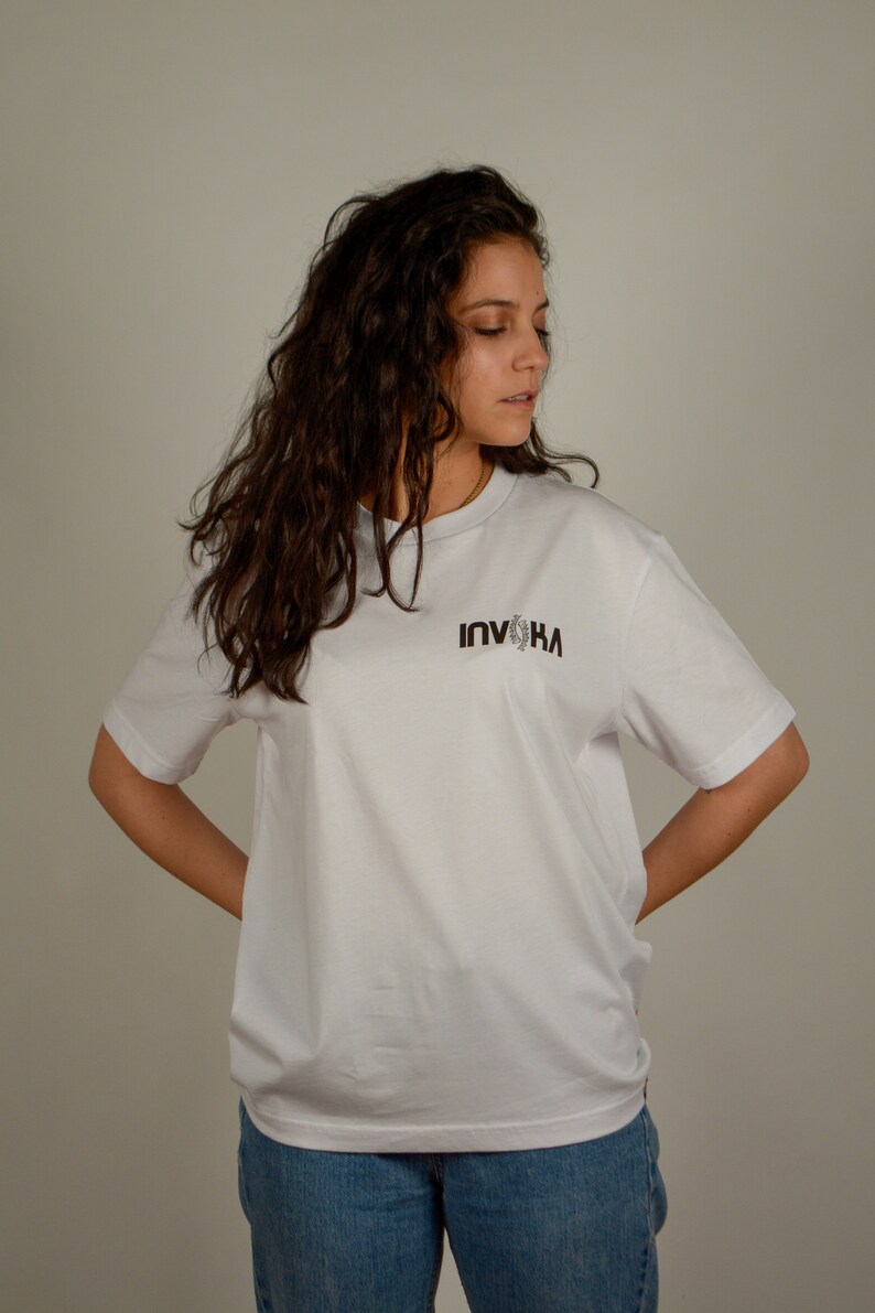 May include: A white t-shirt with a black graphic that says "INVIKA" on the front. The person wearing the shirt has long, curly brown hair.