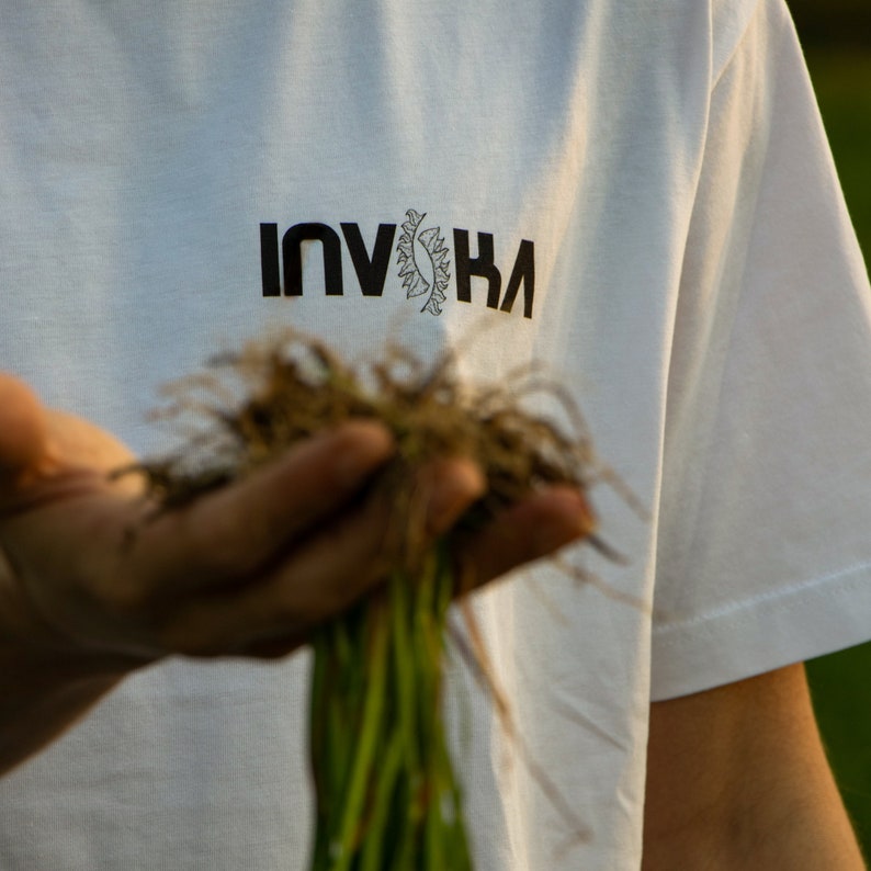 May include: A white t-shirt with the word "INVIKA" printed in black letters. The shirt is being held by a person who is also holding a bunch of green plants.