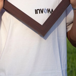 May include: A dark brown wooden picture frame held in front of a white t-shirt. The t-shirt has the word "INVOKA" printed in black with a blue and white design above the word.