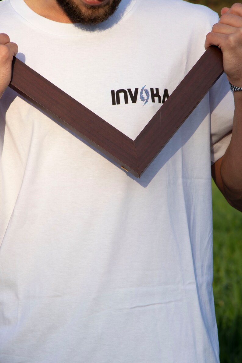 May include: A dark brown wooden picture frame held in front of a white t-shirt. The t-shirt has the word "INVOKA" printed in black with a blue and white design above the word.