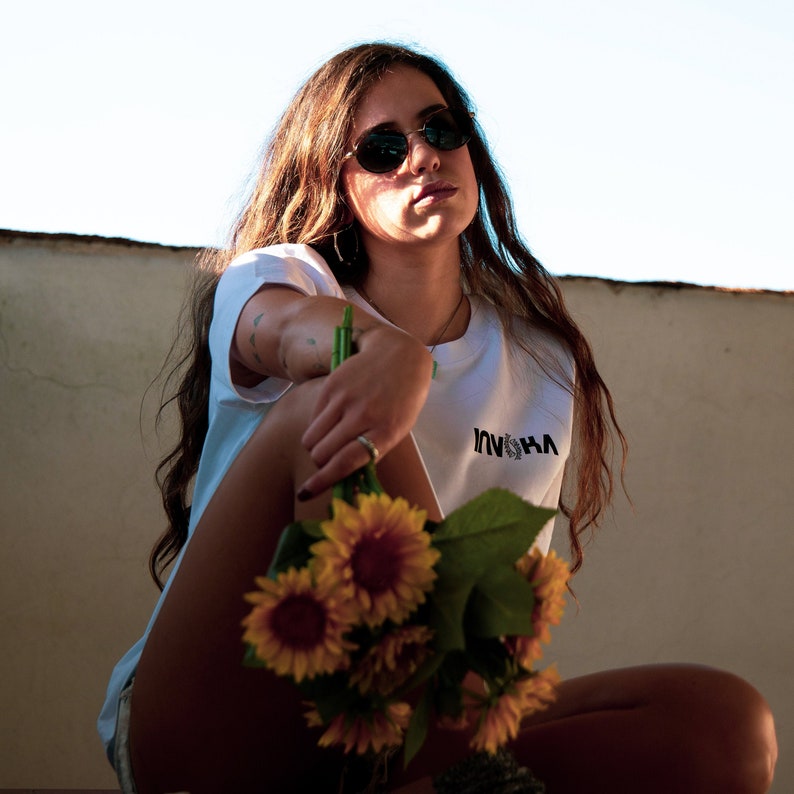 May include: A woman wearing sunglasses and a white t-shirt with the word "INSHA" printed on it, holding a bouquet of yellow sunflowers.
