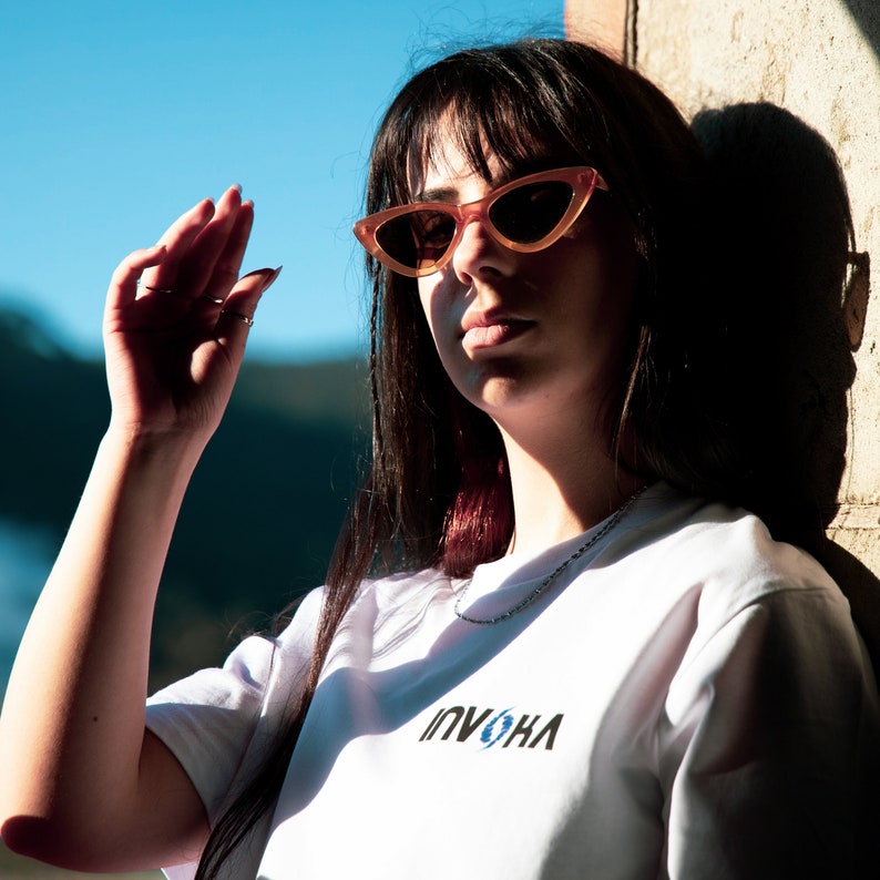 May include: A person wearing orange cat-eye sunglasses and a white t-shirt with the word "INVOKA" printed on it. The person is outside in the sunlight.