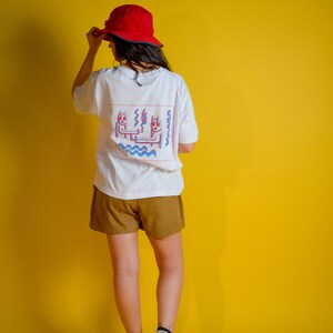 May include: A person wearing a white t-shirt with a colorful graphic on the back, brown shorts, a red hat, and black and white high-top sneakers. The background is a solid yellow.