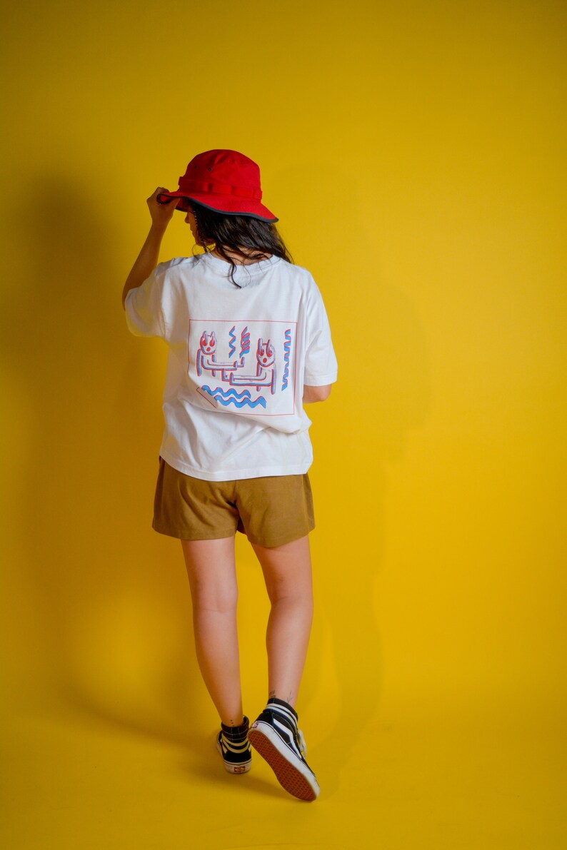 May include: A person wearing a white t-shirt with a colorful graphic on the back, brown shorts, a red hat, and black and white high-top sneakers. The background is a solid yellow.