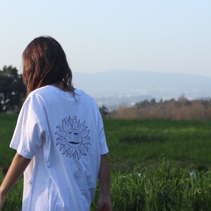 May include: A white t-shirt with a black and white graphic of a smiling sun with a stem and roots. The person wearing the shirt is standing in a field of green grass.