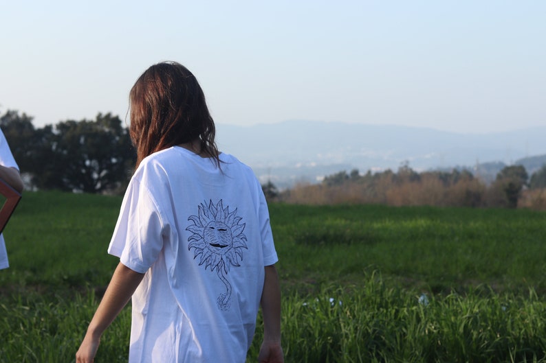 May include: A white t-shirt with a black and white graphic of a smiling sun with a stem and roots. The person wearing the shirt is standing in a field of green grass.