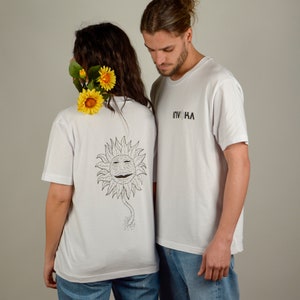 May include: Two people wearing white t-shirts. The person on the left is wearing a t-shirt with a black and white sunflower graphic on the back. The person on the right is wearing a t-shirt with the text "INVOHA" printed on the front. Both people are wearing blue jeans.