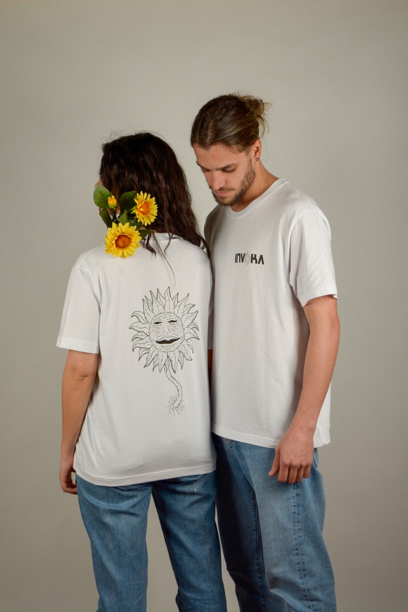 May include: Two people wearing white t-shirts. The person on the left is wearing a t-shirt with a black and white sunflower graphic on the back. The person on the right is wearing a t-shirt with the text "INVOHA" printed on the front. Both people are wearing blue jeans.