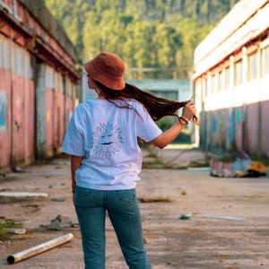 May include: A white t-shirt with a black and white sunflower graphic. The sunflower has a smiling face. The person wearing the shirt is standing in front of a building.