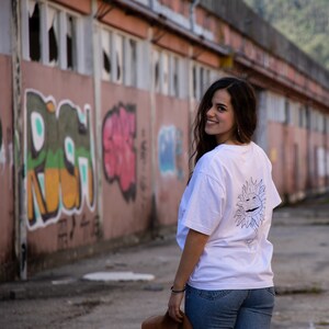 May include: A woman wearing a white t-shirt with a black and white sunflower graphic on the back. She is standing in front of a graffiti-covered wall and looking over her shoulder.