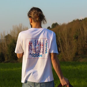 May include: White t-shirt with a colorful graphic on the back. The design features two stylized figures with red and blue accents, set within a red rectangular frame. The figures are holding a wavy line and are above a blue wavy line.