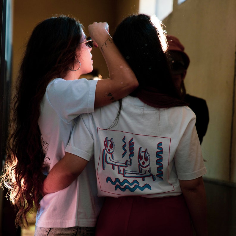 May include: Two people wearing white t-shirts with a graphic design on the back, embracing each other. The graphic features two figures with red and blue accents, and wavy lines. One person wears sunglasses and the other wears a red skirt.