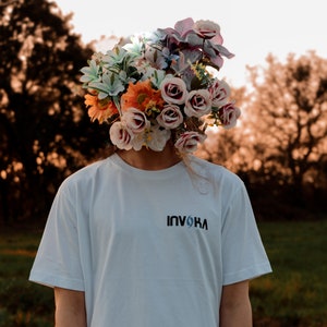 May include: A person wearing a white t-shirt with the word "INVOKA" printed on the front. The person's head is replaced by a large arrangement of colorful flowers, including roses and lilies, against a blurred outdoor background.