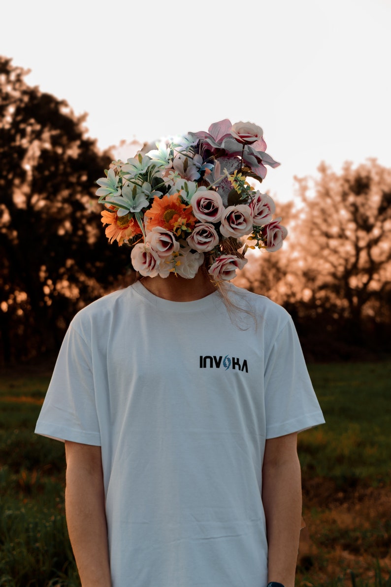 May include: A person wearing a white t-shirt with the word "INVOKA" printed on the front. The person's head is replaced by a large arrangement of colorful flowers, including roses and lilies, against a blurred outdoor background.