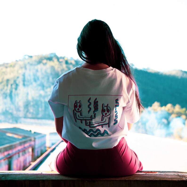 May include: White t-shirt with a graphic design on the back featuring two cartoon figures with wavy lines and the word "Wawa" in teal. The person is wearing maroon pants and is sitting on a wooden ledge.