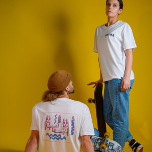 May include: Two people wearing white t-shirts with the word "RVOKA" in blue. One shirt has a graphic on the back. They are posing with skateboards against a yellow background. The person in the foreground is wearing a brown beanie.