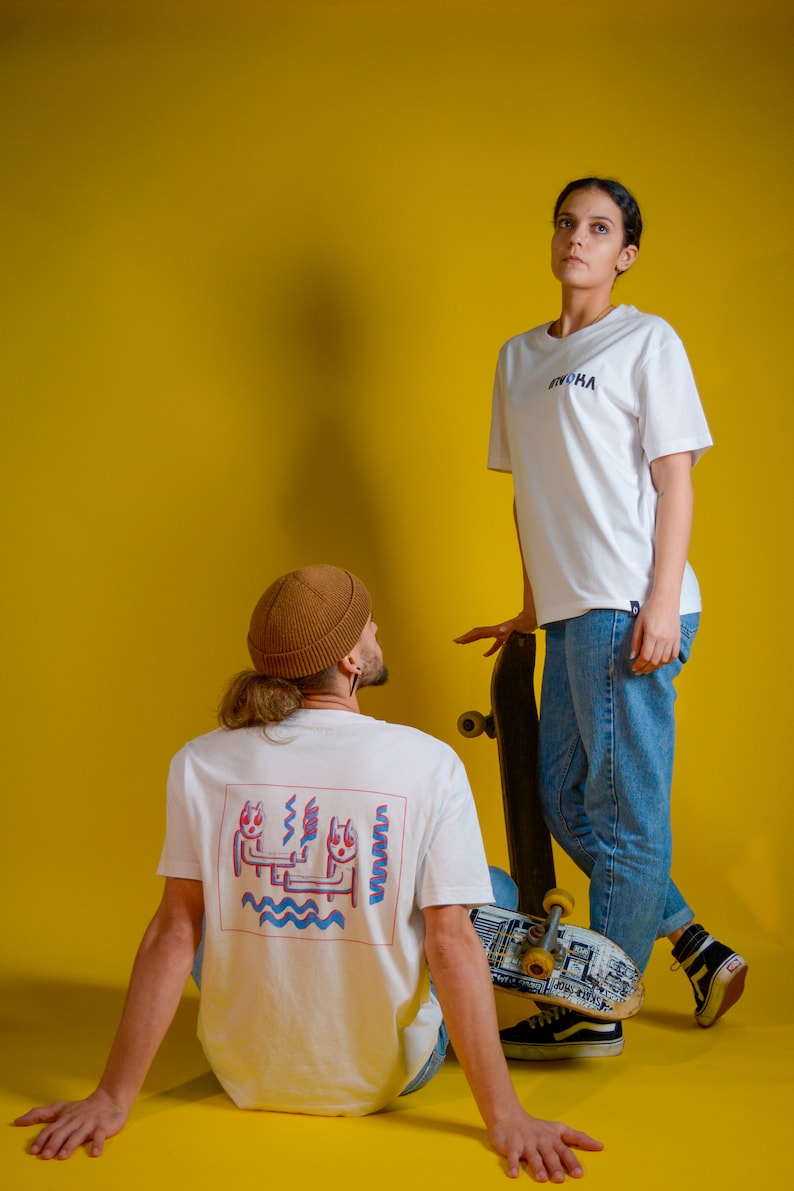 May include: Two people wearing white t-shirts with the word "RVOKA" in blue. One shirt has a graphic on the back. They are posing with skateboards against a yellow background. The person in the foreground is wearing a brown beanie.